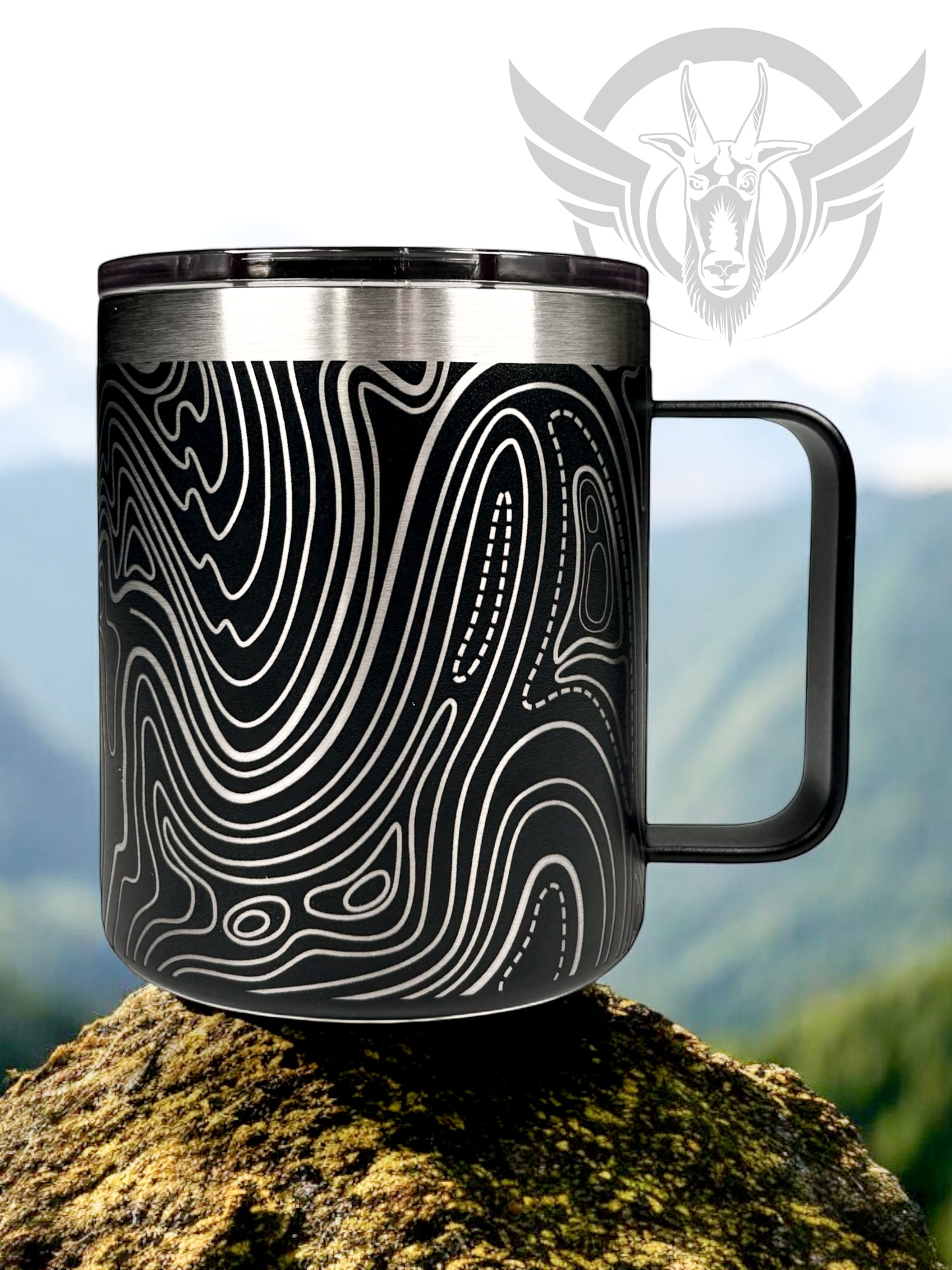 A black travel mug with a handle features a wrap-around line topography design. The cup sits on a rock in a natural outdoor setting.
