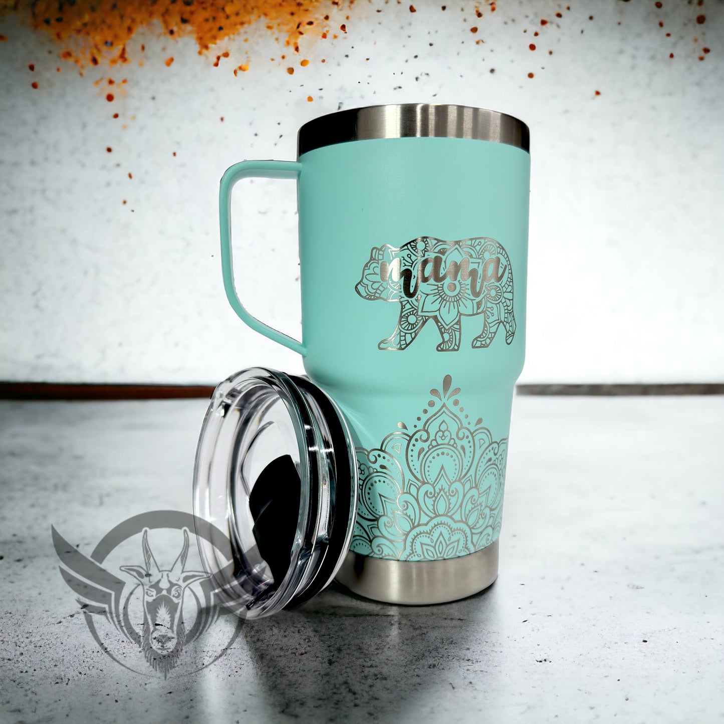 A light teal travel mug with a lid beside it and handle show off an intricate mandala design along the bottom front. Above the design is a bear shaped image made with a mandala design and includes the word "mama" within it. The cup is displayed on a concrete surface.