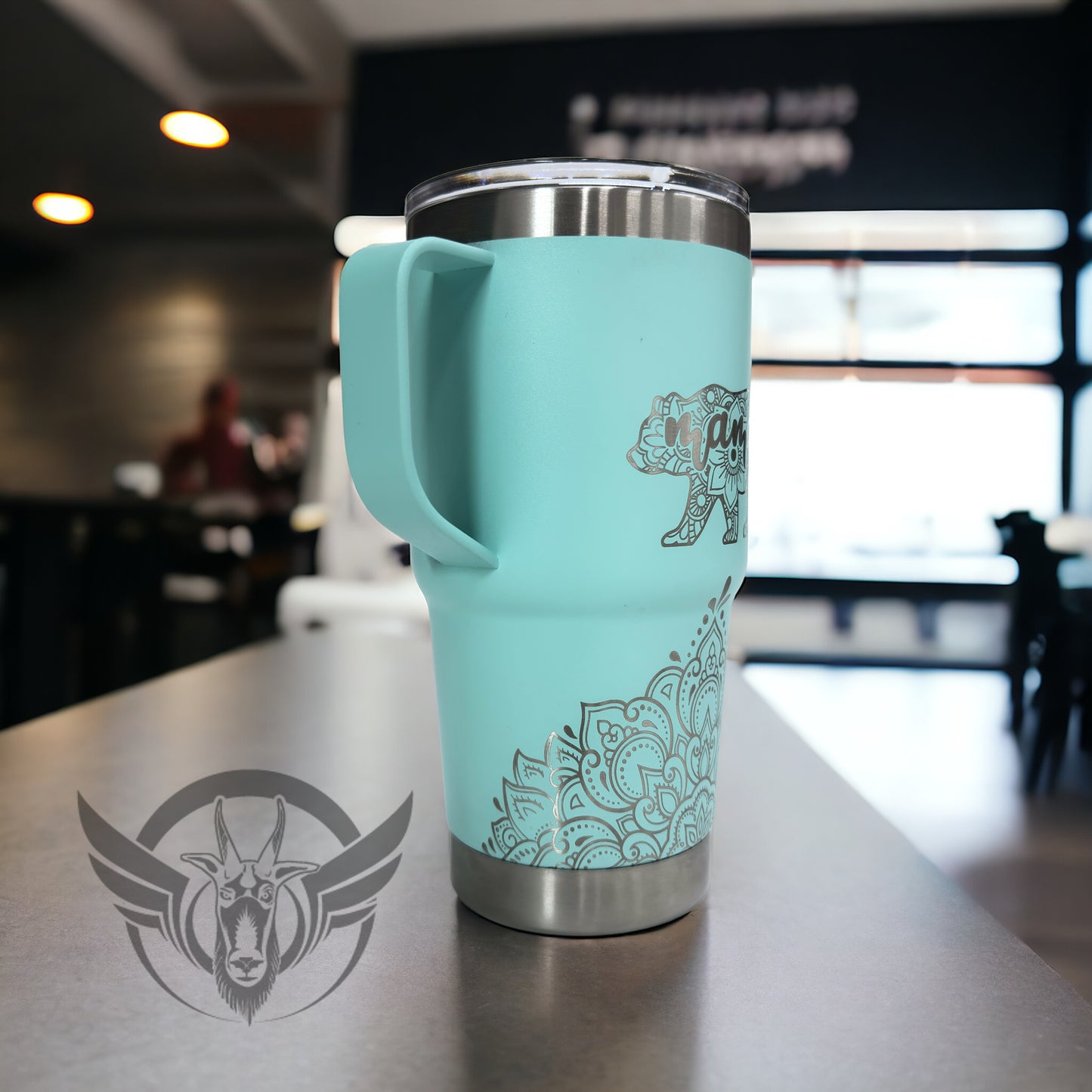 A light teal travel mug with a lid and handle show off an intricate mandala design along the bottom front, the cup is at an angle to show the left side of it. Above the design is a bear shaped image made with a mandala design and includes the word "mama" within it. The cup is displayed on a bar top .