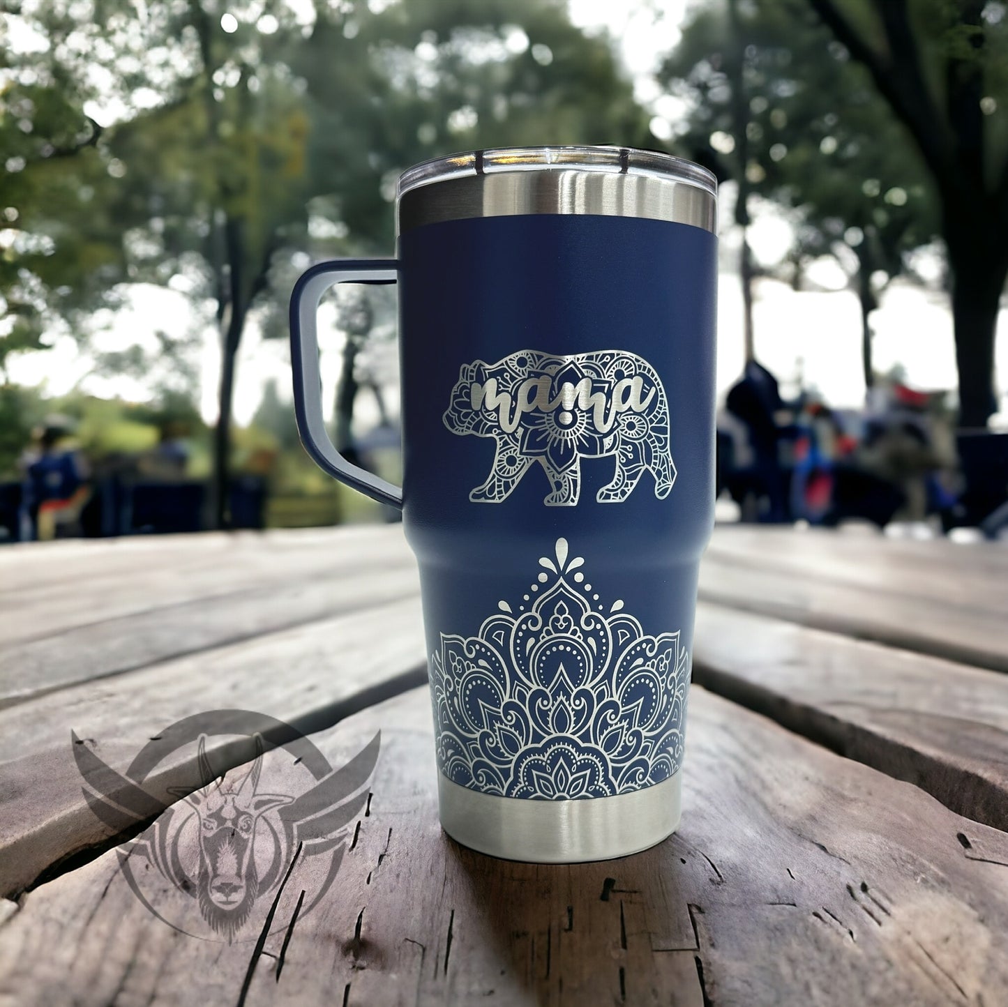 A navy blue travel mug with a lid and handle show off an intricate mandala design along the bottom front. Above the design is a bear shaped image made with a mandala design and includes the word "mama" within it. The cup is displayed on a wooden surface outdoors.